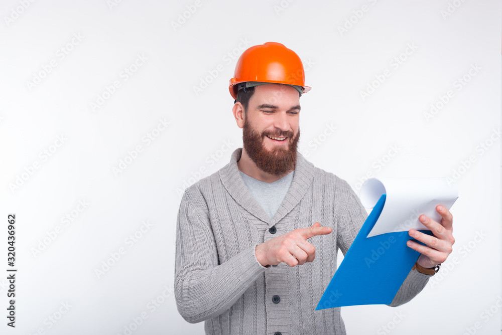 Young constructor man is checking his list. He wears an orange hard protection hat.