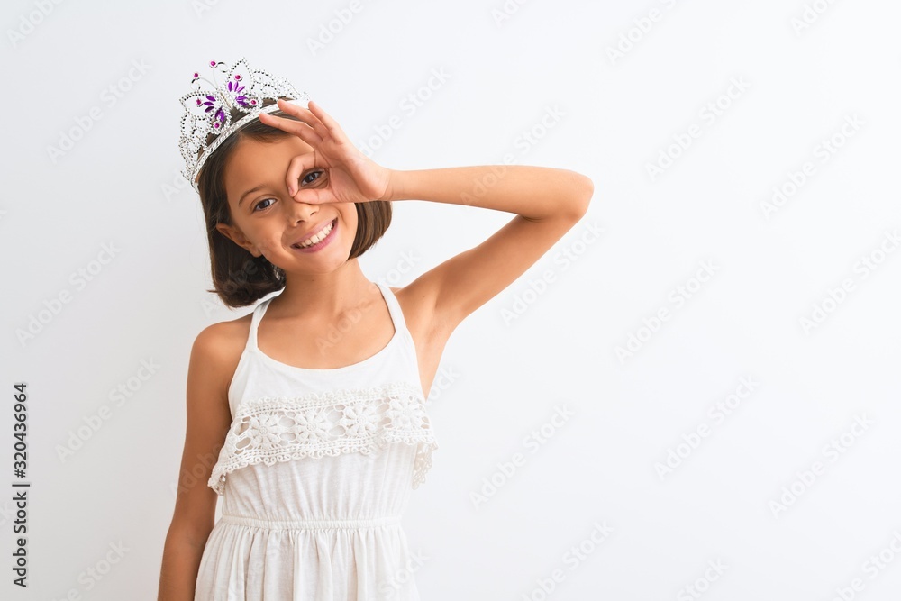 Beautiful child girl wearing princess crown standing over isolated white background doing ok gesture with hand smiling, eye looking through fingers with happy face. Beautiful child girl wearing princess crown standing over isolated white background doing ok gesture with hand smiling, eye looking through fingers with happy face.