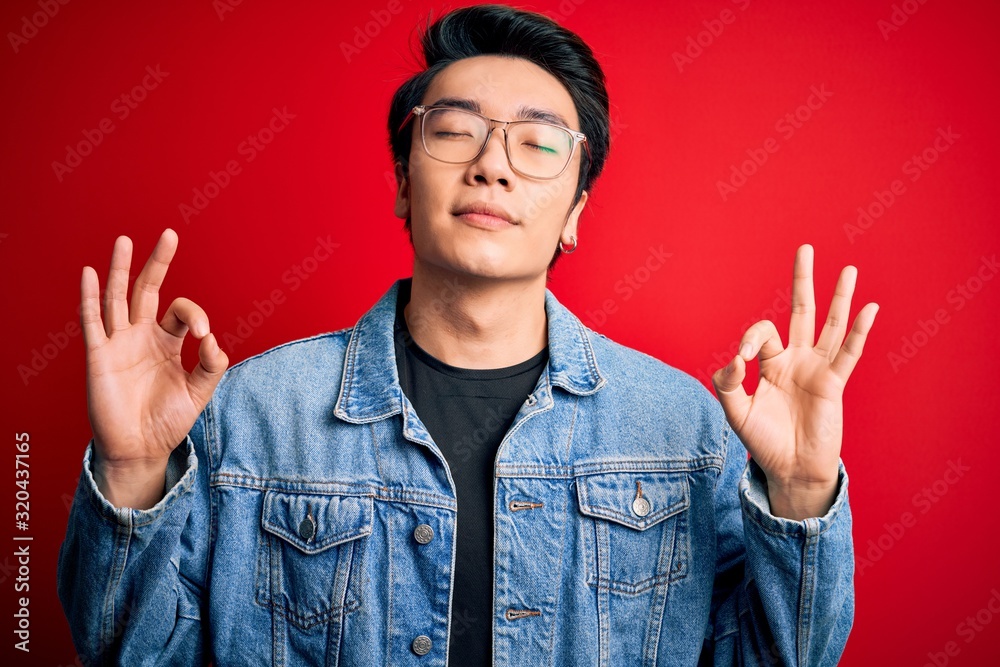 Young handsome chinese man wearing denim jacket and glasses over red background relax and smiling with eyes closed doing meditation gesture with fingers. Yoga concept. Young handsome chinese man wearing denim jacket and glasses over red background relax and smiling with eyes closed doing meditation gesture with fingers. Yoga concept.