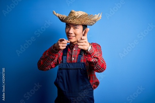 Young handsome chinese farmer man wearing apron and straw hat over blue background pointing fingers to camera with happy and funny face. Good energy and vibes.