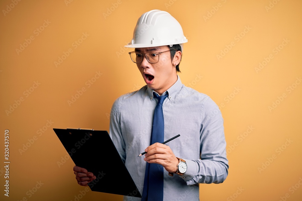 Young handsome chinese engineer man wearing safety helmet holding clipboard scared in shock with a surprise face, afraid and excited with fear expression Young handsome chinese engineer man wearing safety helmet holding clipboard scared in shock with a surprise face, afraid and excited with fear expression