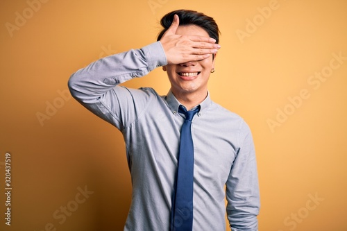 Young handsome chinese businessman wearing glasses and tie over yellow background smiling and laughing with hand on face covering eyes for surprise. Blind concept.