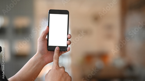 Man's hand holding crop black smartphone with white blank screen and pointing finger on button with modern living room as blurred background.
