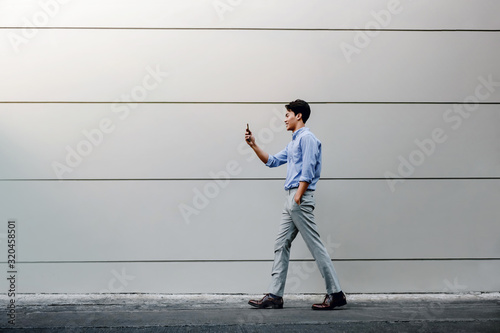 Happy Young Businessman in Casual wear Using Mobile Phone while Walking by the Urban Building Wall. Lifestyle of Modern People. Side View. Full Length