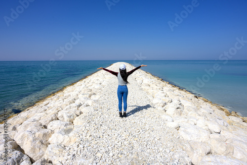 Woman standing on the pier white rocks showing hands up to blue sky over sea water background, Bahrain.