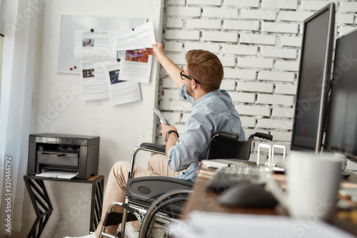 Working day. Young busy male office worker in a wheelchair putting some documents on white board while sitting near printer at his workplace in the bright modern office