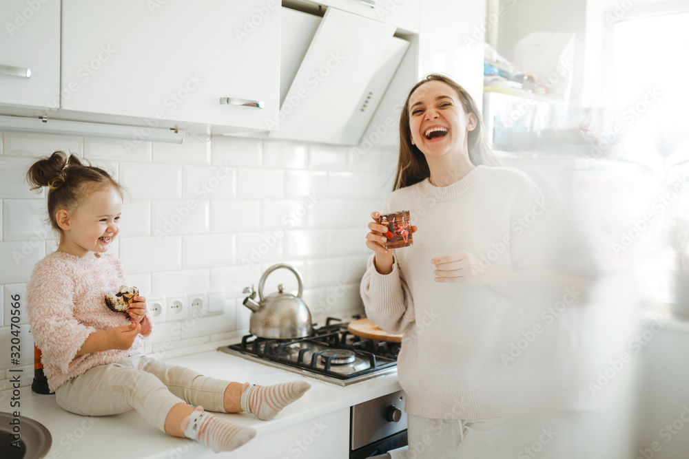Mother and little daughter have fun together in the sunny kitchen. Little girl sitting on table top and eats a bakery products with mom. Happy loving family. Mother and little daughter have fun together in the sunny kitchen. Little girl sitting on table top and eats a bakery products with mom. Happy loving family.