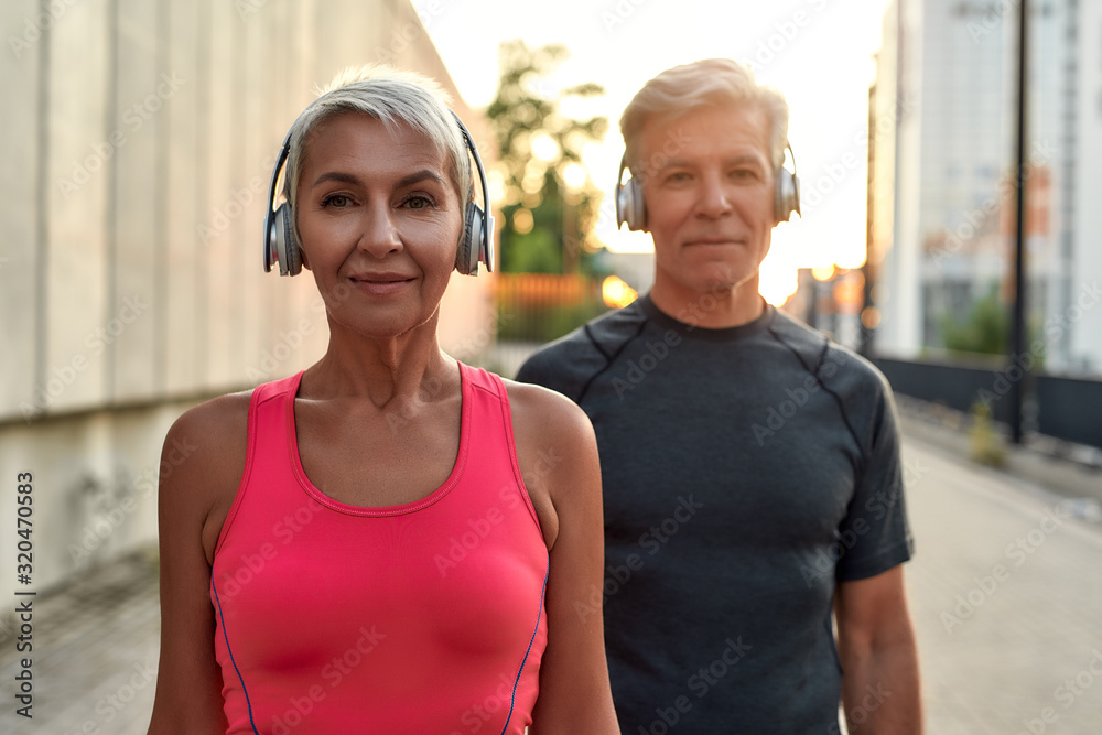 Training with music. Portrait of happy and beautiful middle-aged couple in headphones looking at camera with smile while training together outdoors