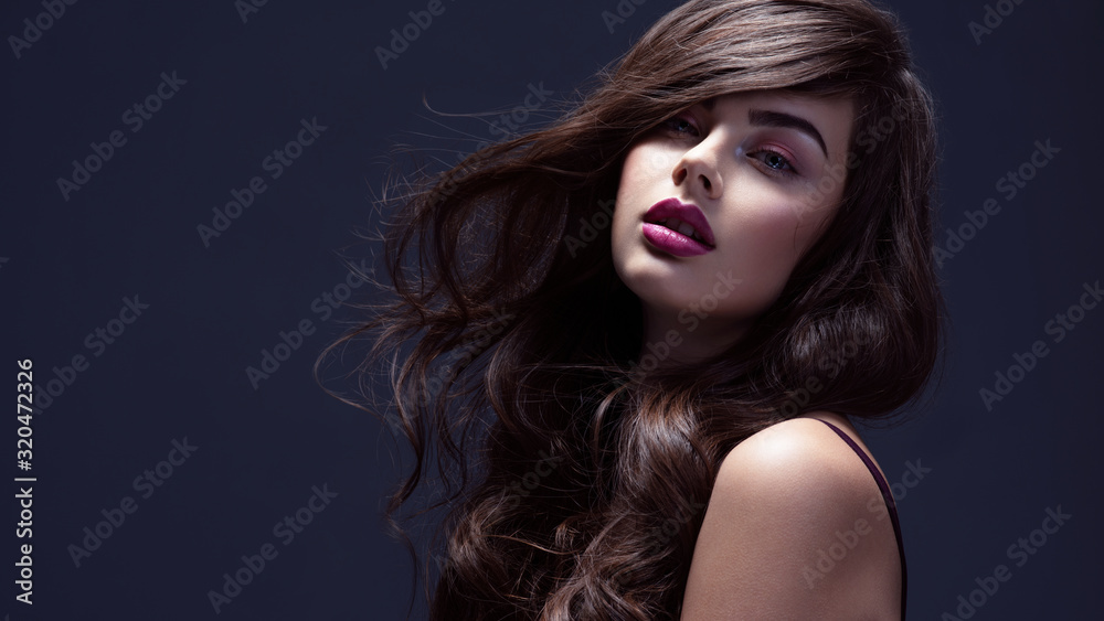 Naklejka premium Face of a beautiful woman with long brown curly hair. Fashion model with wavy hairstyle. Attractive young girl with curly hair posing at studio. Female face with purple makeup. Violet make-up.