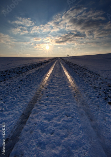 Eisige Strasse mit Reifenspuren im Schnee auf denen sich die Abendsonne auf dem Eis spiegelt