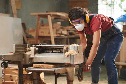 Concentrated female carpenter in mask, safety glasses and earmuffs processing wooden board on woodworking machine