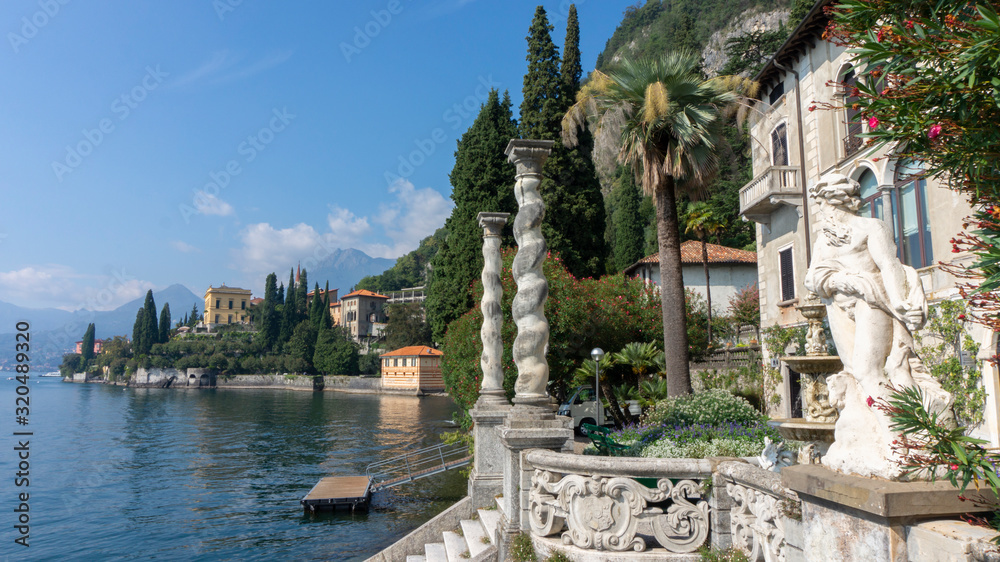 Scenic view of the Monastero villa with antique statues, front columns and a marble stair to the pier, surrounded by palms and thuja. View of an antique European city on the shore of an alpine lake