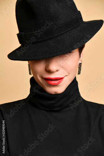 Portrait of young smiling woman in black hat worn to one side, black poloneck and earings over dark yellow wall background photo