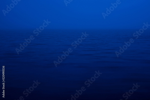 Fototapeta Naklejka Na Ścianę i Meble -  Natural texture of deep blue calm water in dusk close up. Night sea of blue classic color. Water ripple nature background. Meditative image of thick fog above lake. Soft light shines on water surface.