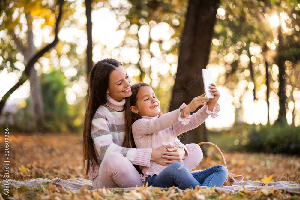 Mother and daughter using smartphone in autumn in park. - obrazy, fototapety, plakaty 