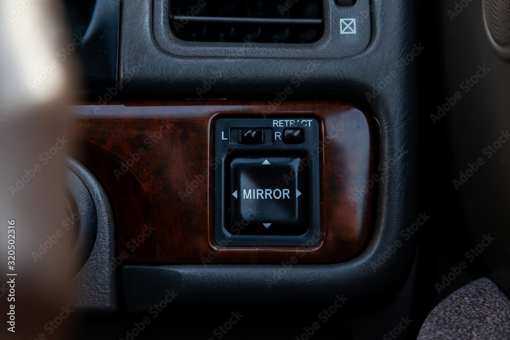 The buttons for controlling the mirrors electric drive on a stylish panel covered with wood inside the car are of modern design in black with white signs and symbols.