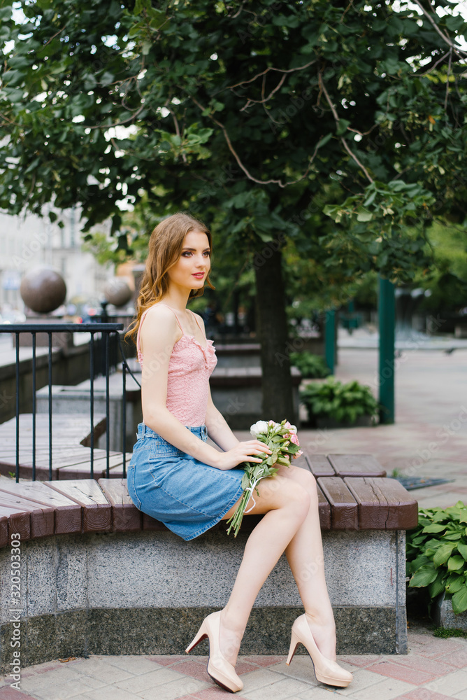 A beautiful romantic girl in a pink top and denim skirt holding a bouquet of roses and sitting on a bench in the city square A beautiful romantic girl in a pink top and denim skirt holding a bouquet of roses and sitting on a bench in the city square