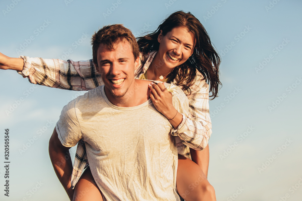 Fototapeta premium Handsome young man giving piggyback ride to girlfriend on beach. Young couple enjoying summer holidays.