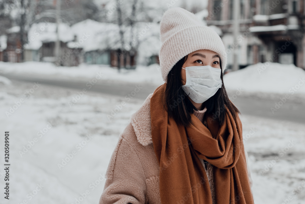 Plakat Passerby Asian girl in a protective medical mask outdoors. A woman on the street in winter protects the respiratory tract from the epidemic of coronavirus. Protection against urban smog and dust