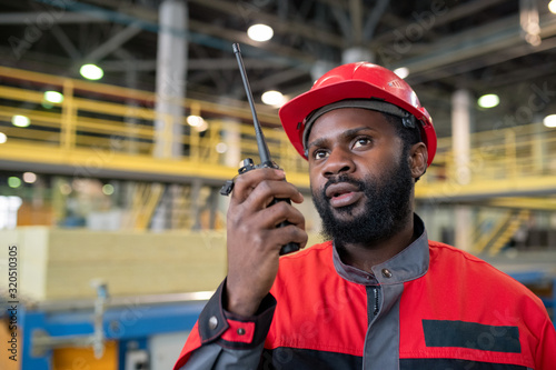 Young bearded Black laborer in red uniform transferring message to coworker with walkie-talkie device at construction site