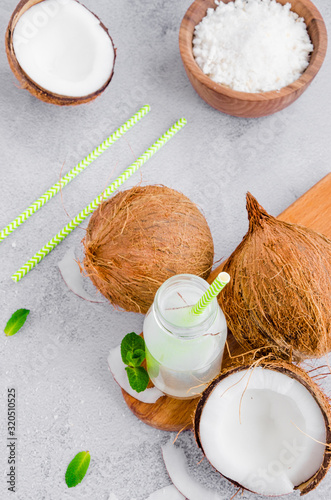 Fresh cold coconut water in a glass bottle with a straw and broken coconuts on a gray background. A healthy drink concept. Vertical orientation. Copy space