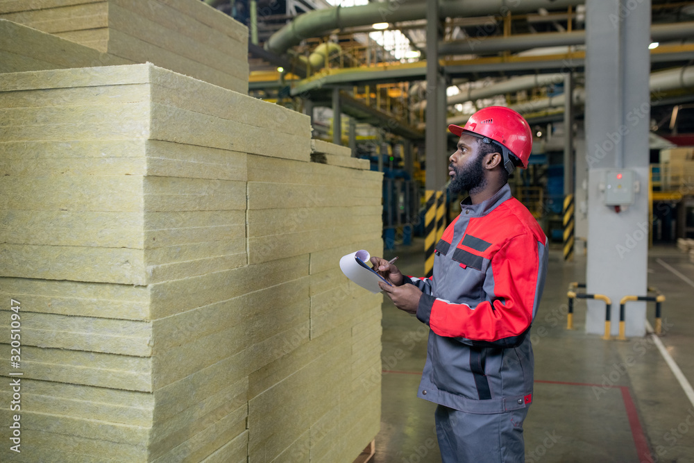 Serious young bearded Black builder counting produced heat materials and making notes in clipboard Serious young bearded Black builder counting produced heat materials and making notes in clipboard