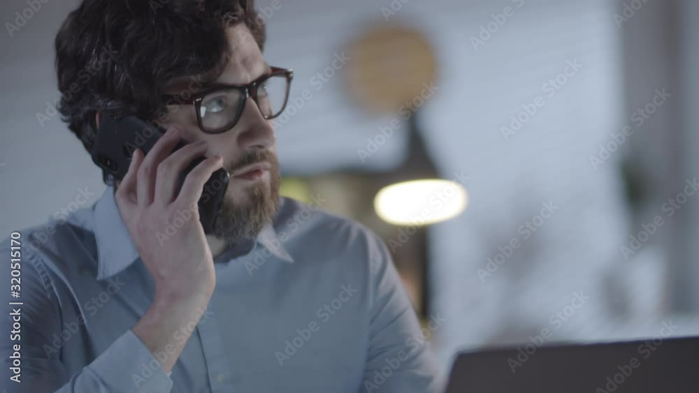 Tilt up shot of bearded businessman in shirt and glasses sitting at his desk and talking on phone while working in office in evening Tilt up shot of bearded businessman in shirt and glasses sitting at his desk and talking on phone while working in office in evening