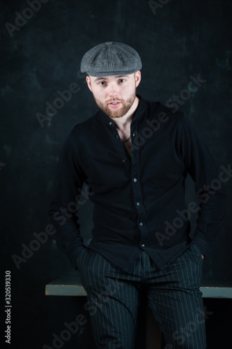 Serious handsome young bearded man in hat on grey background looking at camera. Portrait of man against grey wall.