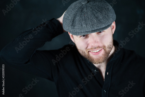 Handsome young bearded man in hat on grey background looking at camera. Portrait of laughing young man against grey wall. Happy guy smiling.