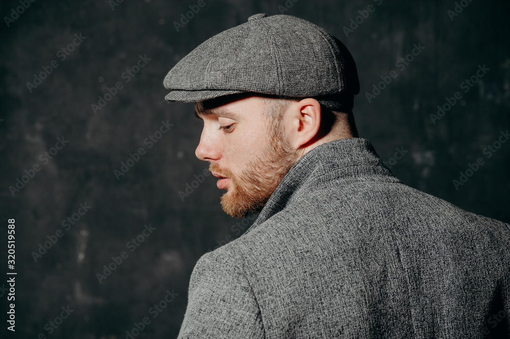 Serious handsome young bearded man in hat on grey background looking at side. Portrait of man against grey wall. Serious handsome young bearded man in hat on grey background looking at side. Portrait of man against grey wall.