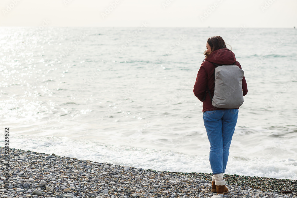 A woman in a jacket with a backpack watches the sea waves view from behind.