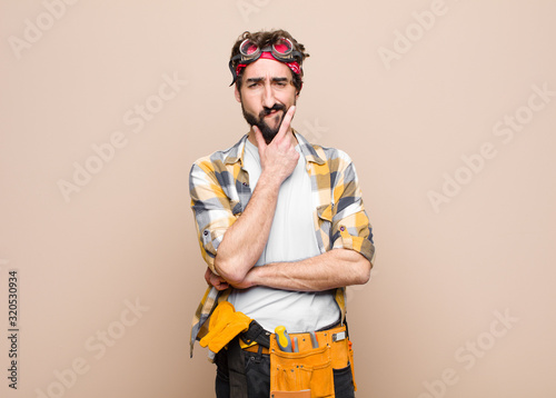 young housekeeper man looking serious, thoughtful and distrustful, with one arm crossed and hand on chin, weighting options against flat wall photo