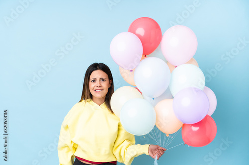 Woman holding balloons in a party over isolated blue background having doubts and with confuse face expression © luismolinero