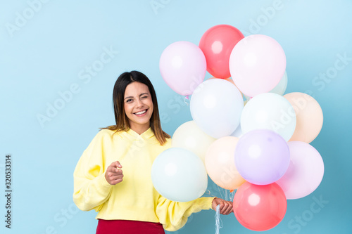 Woman holding balloons in a party over isolated blue background points finger at you