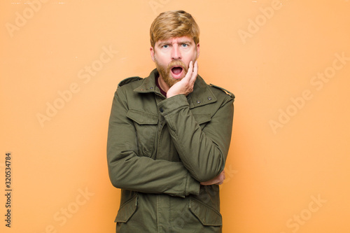 young blonde man open-mouthed in shock and disbelief, with hand on cheek and arm crossed, feeling stupefied and amazed against flat wall photo