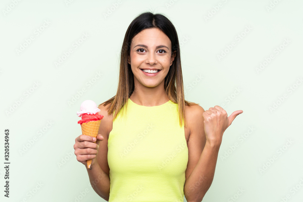 Brunette woman holding a cornet ice cream over isolated green background pointing to the side to present a product Brunette woman holding a cornet ice cream over isolated green background pointing to the side to present a product