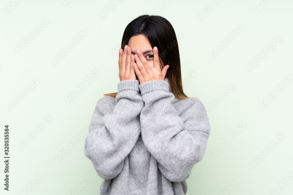 Woman wearing a sweater over isolated green background covering eyes and looking through fingers Woman wearing a sweater over isolated green background covering eyes and looking through fingers