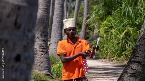 Young black man in a orange shirt tying a scarf on his hand while walking between palm trees with a hat in Tanzania