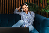 Young hipster girl sitting on sofa in white headphones and looking at laptop screen. Young woman relaxing on couch watching online movies, streaming services.Beautiful girl smiling, looking at came.