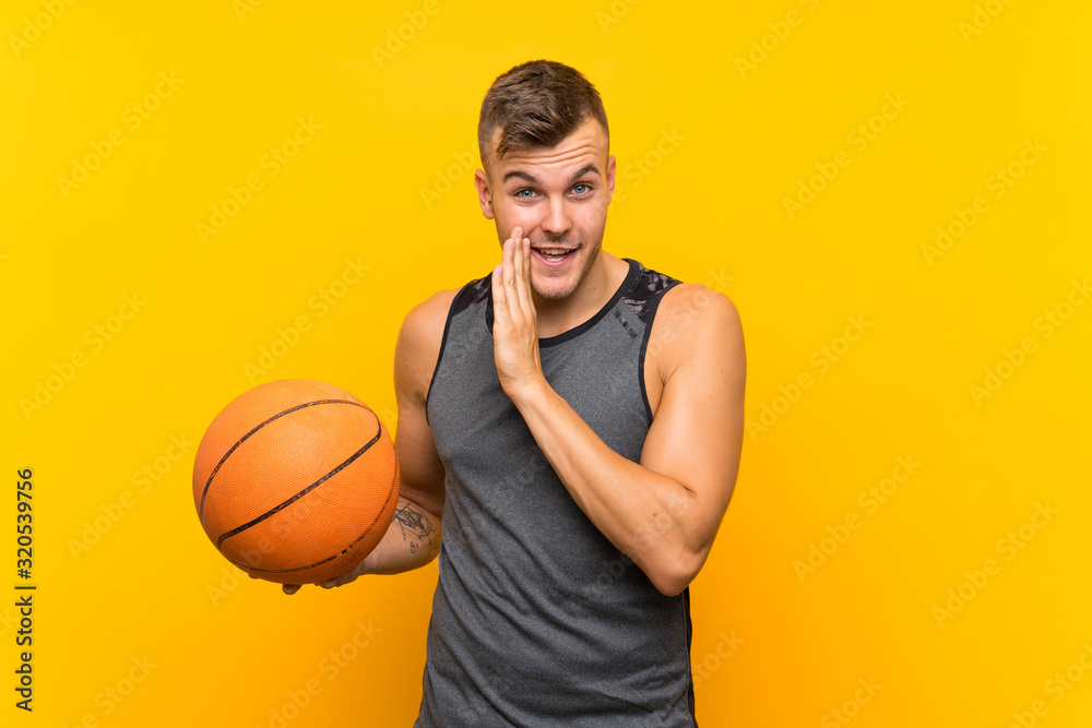 Young handsome blonde man holding a basket ball over isolated yellow background whispering something