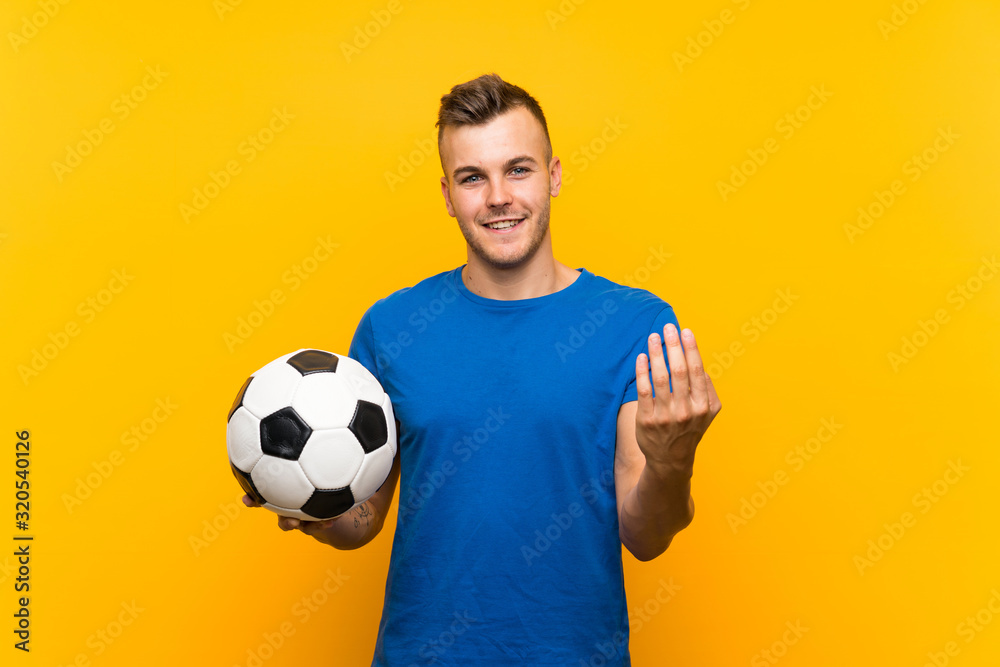 Young handsome blonde man holding a soccer ball over isolated yellow background inviting to come with hand. Happy that you came Young handsome blonde man holding a soccer ball over isolated yellow background inviting to come with hand. Happy that you came