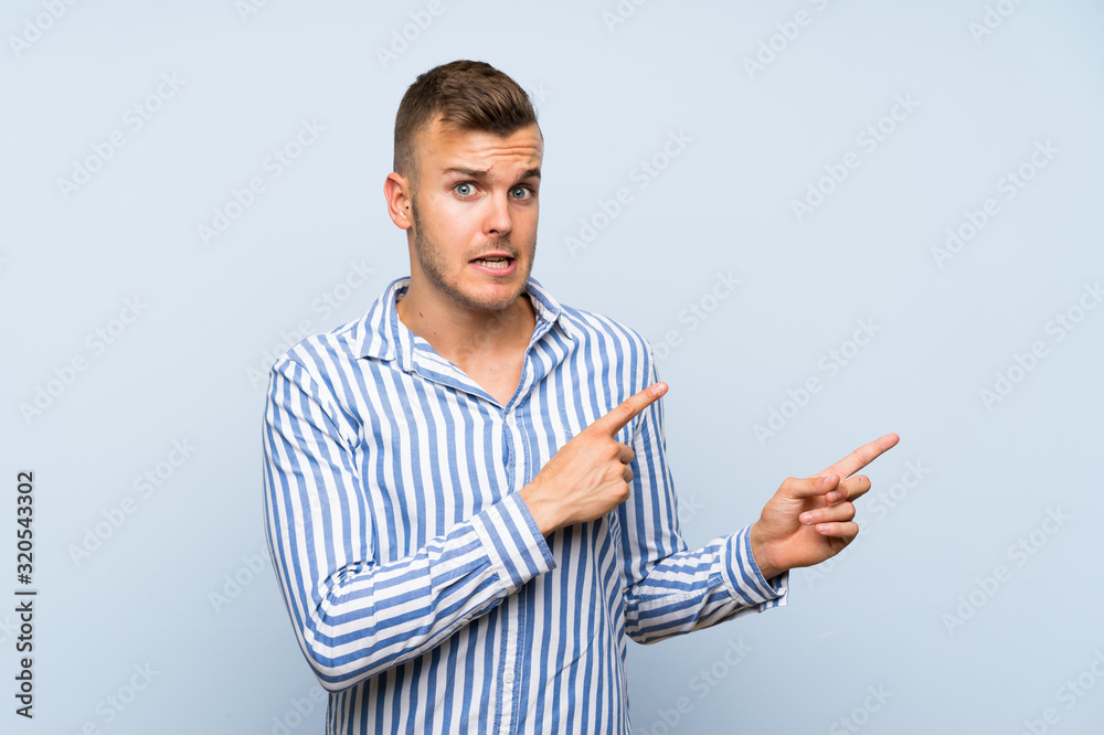 Young handsome blonde man over isolated blue background frightened and pointing to the side Young handsome blonde man over isolated blue background frightened and pointing to the side