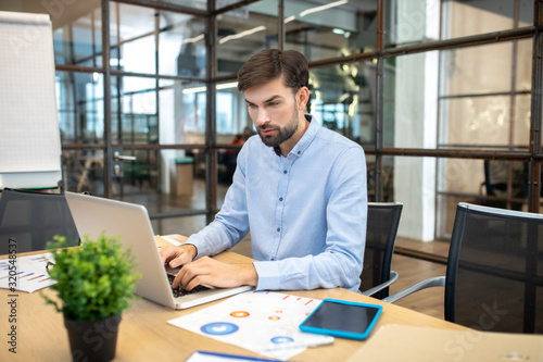 Bearded man in a blue shirt sitting in the office and working on laptop