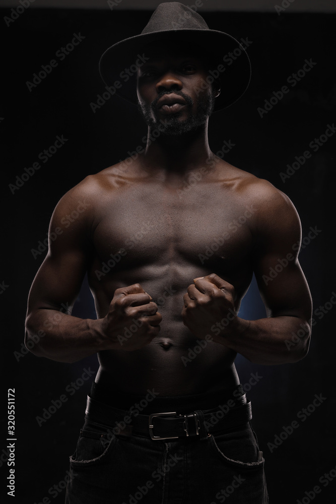 portrait of a handsome black man in dark hat and jeans with naked sports torso looking in the camera on dark background and holds his fists in front of him