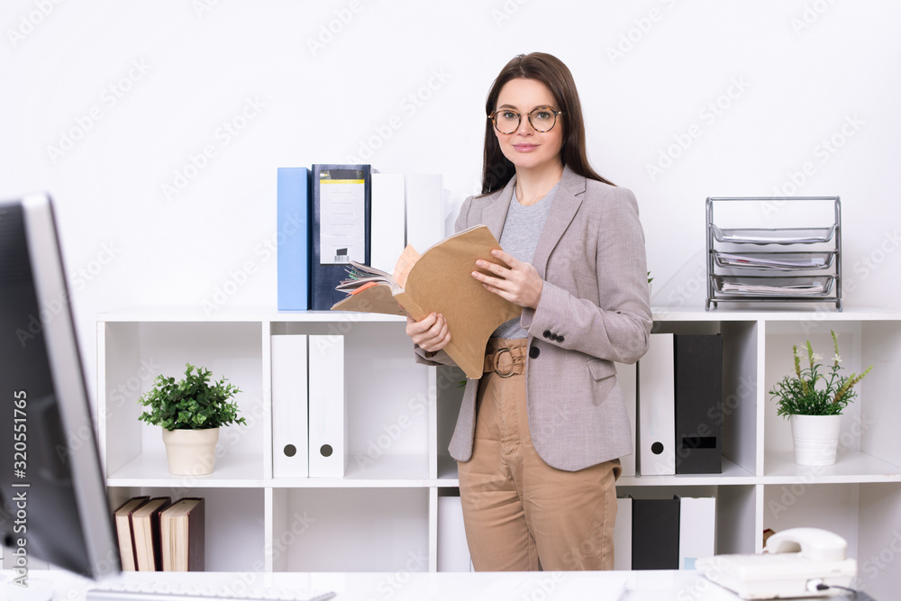 Portrait of content stylish secretary in eyeglasses standing at office shelves and finding file in folder