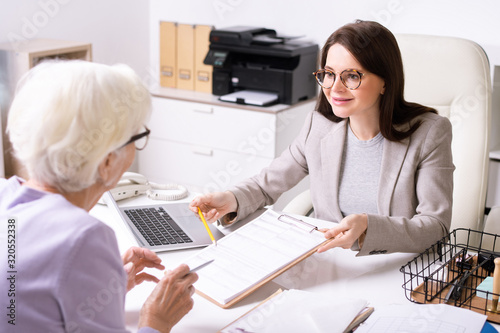 Smiling attractive young woman in glasses sitting at desk and pointing at paper while explaining senior lady how to fill form