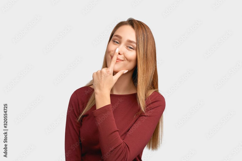 Funny girl holding index finger on nose, smiling sweetly. Beautiful young woman with long hair, clean smooth skin isolated on white background in Studio with an empty space for text and advertising. Funny girl holding index finger on nose, smiling sweetly. Beautiful young woman with long hair, clean smooth skin isolated on white background in Studio with an empty space for text and advertising.