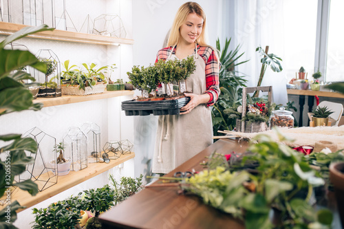 careful florist lady with long blonde hair in red casual checkered shirt and apron stand in her own florist shop, look after plants and flowers