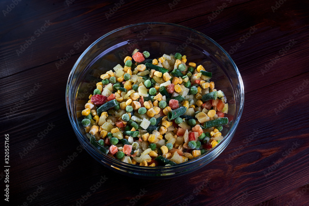 Frozen different vegetables in a glass plate on a wooden background. - obrazy, fototapety, plakaty 