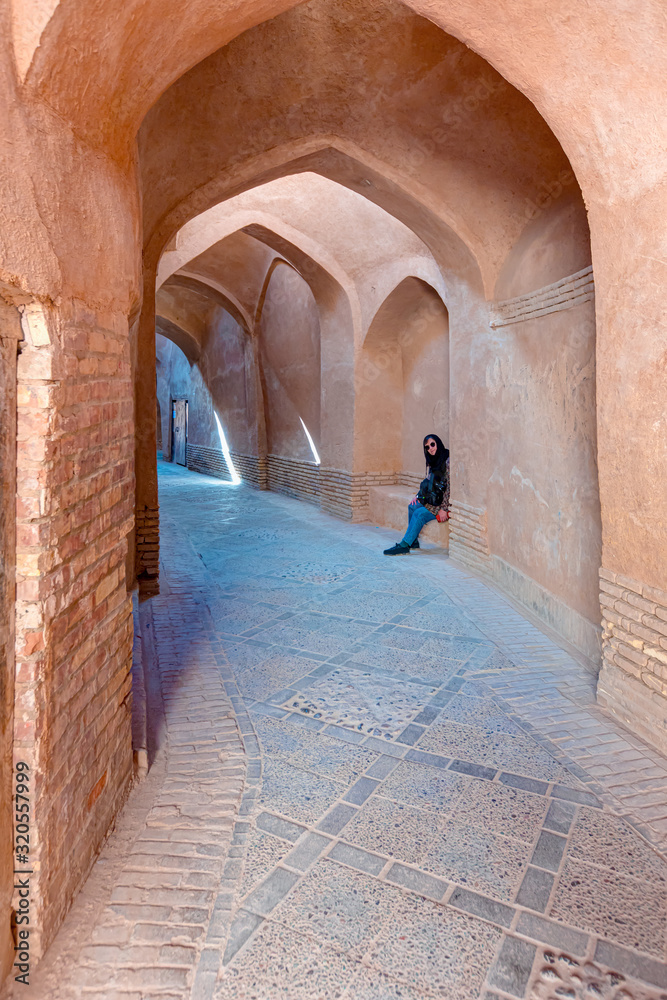 Female tourist resting on the old antique brick wall - High narrow street of old adobe town with famous wind towers - Yazd, Iran
